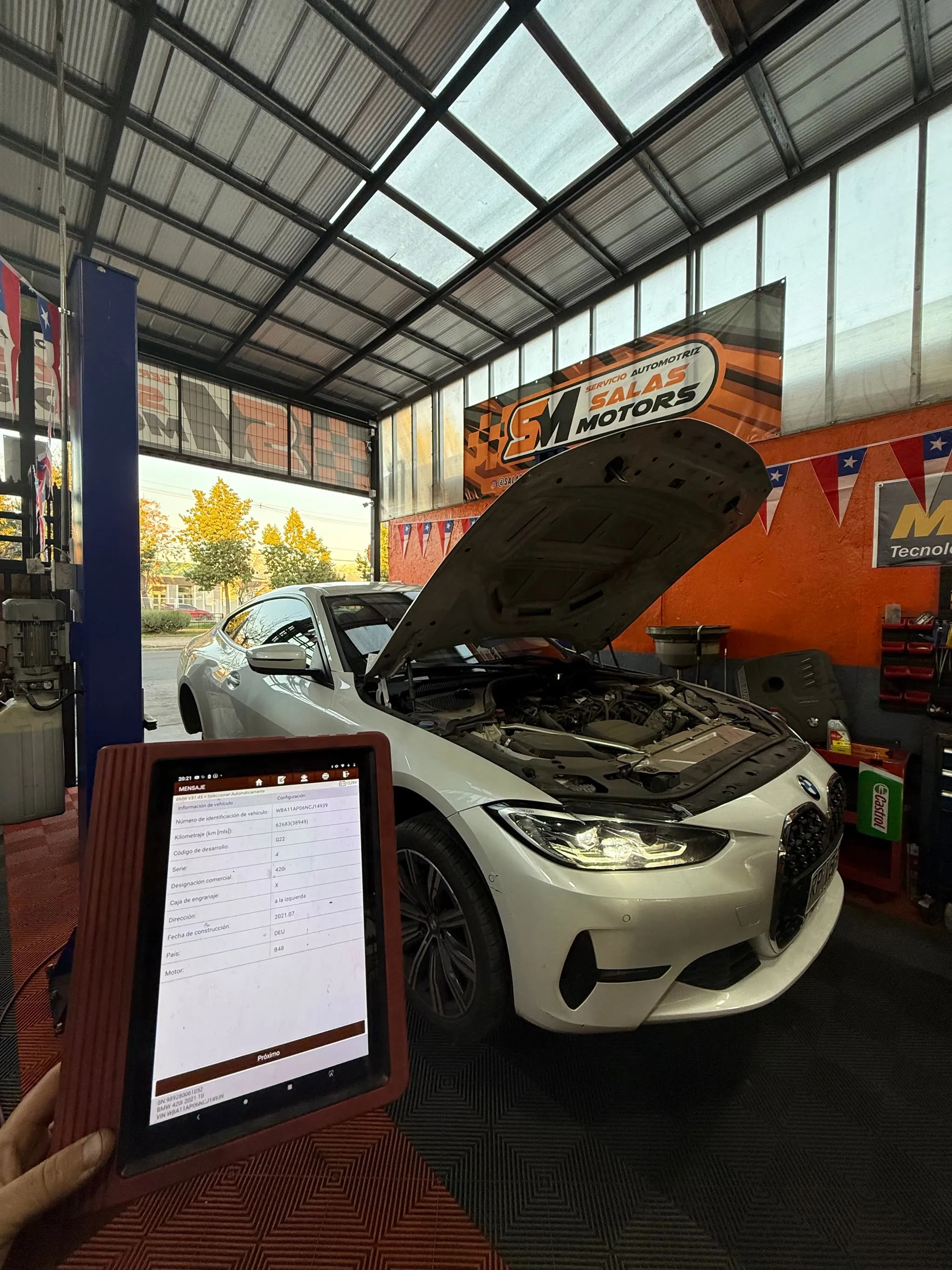 Scanner de diagnóstico en la mano de un técnico, frente a un BMW blanco con el capó abierto en el taller